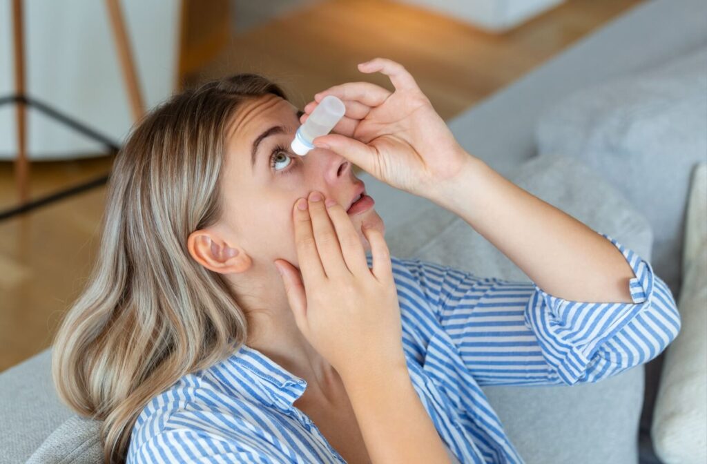 A person sitting indoors tilts their head back while applying eye drops, using one hand to hold the bottle above their eye and the other to gently pull down their lower eyelid.